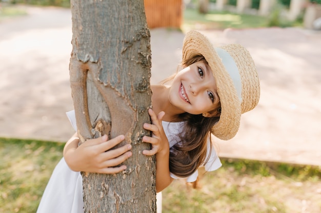 Free photo funny dark-haired kid with big eyes and smile embracing tree in park. outdoor portrait of happy little girl in straw hat enjoying summer vacation.