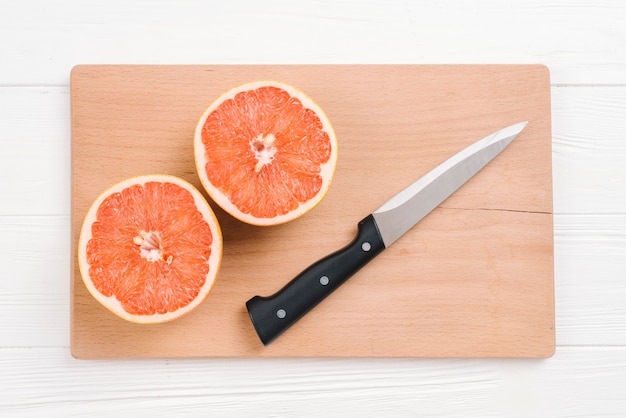 Free photo halved grapefruits with sharp knife on wooden chopping board over white desk