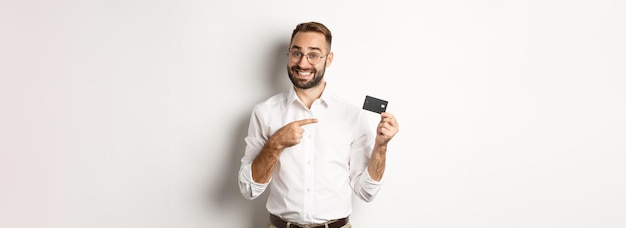 Free Photo handsome satisfied man in glasses pointing at credit card pleased with bank services standing over w