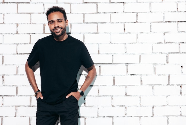 Free photo handsome smiling hipster model sexy unshaven man dressed in black summer tshirt and jeans clothes fashion male posing near white brick wall in studio at sunny day isolated