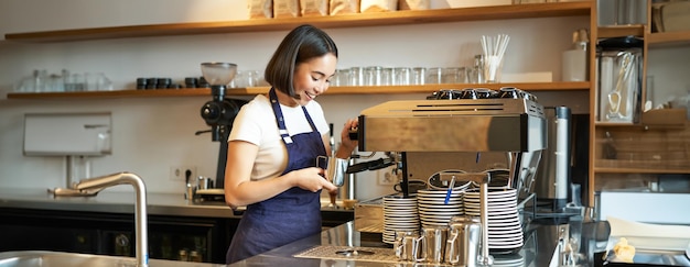 Free photo happy asian girl barista working in cafe brewing coffee prepare cappuccino using steamer on machine