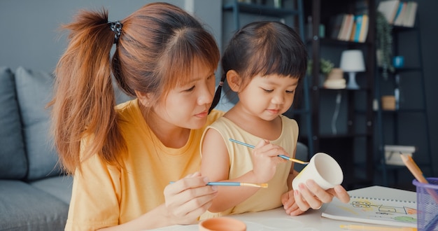 Free Photo happy cheerful asia family mom teach toddler girl paint ceramic pot having fun relax on table in living room at house