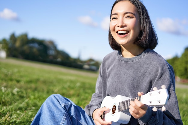 Free photo happy people and hobbies smiling asian girl playing ukulele guitar and singing sitting in park outdo