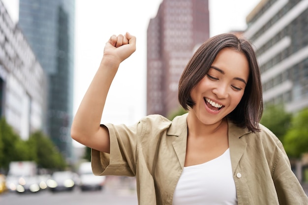 Free photo happy people young smiling asian woman dancing in city raising hands up in city triumphing celebrati