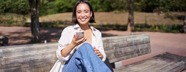 Free Photo happy smiling asian girl sitting on bench in park and browsing on social media holding smartphone