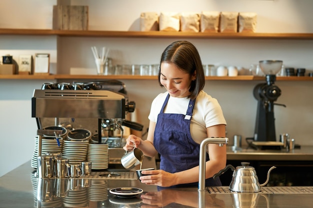 Free photo happy smiling cafe owner girl barista in apron making cappuccino latte art with steamed milk standin