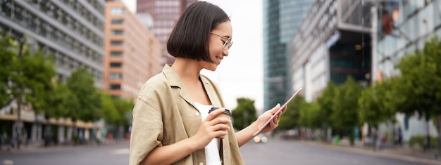 Free Photo happy woman exploring the city young korean girl holds tablet drinks coffee and walks along street