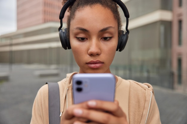 Free photo horizontal shot of sporty woman listens music via headphones holds mobile phone concentrated into screen wears hoodie rests after fitness training rests after practicing yoga leads active lifestyle