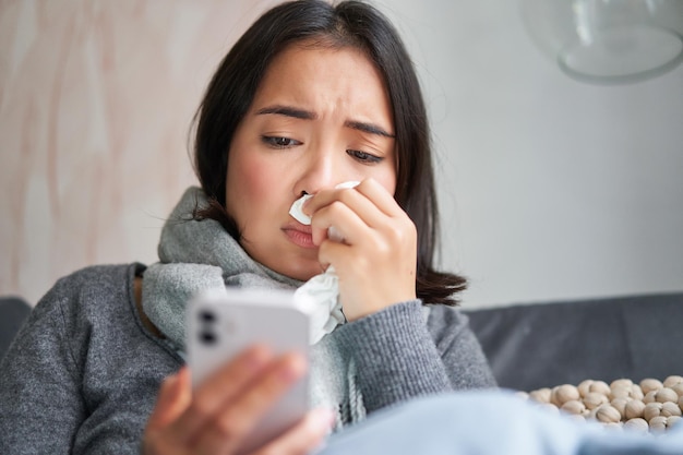 Free Photo ill korean woman sitting at home sneezing caught cold reading news on mobile phone freezing at her h