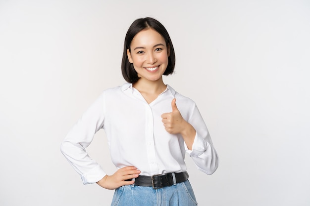 Free photo image of confident asian woman showing thumb up in approval recommending like smth good standing over white background