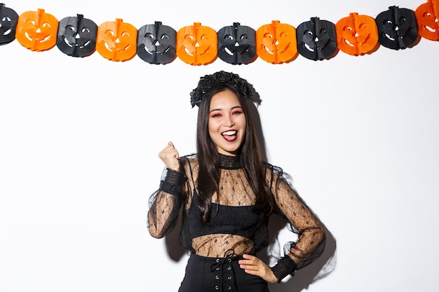 Image of happy asian woman in witch costume having fun on halloween party, saying yes cheerful, standing against pumpkin banners.