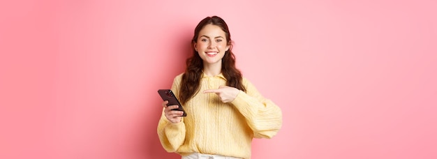 Image of happy smiling young woman holding mobile phone and pointing at mobile screen with pleased and confident face standing over pink background