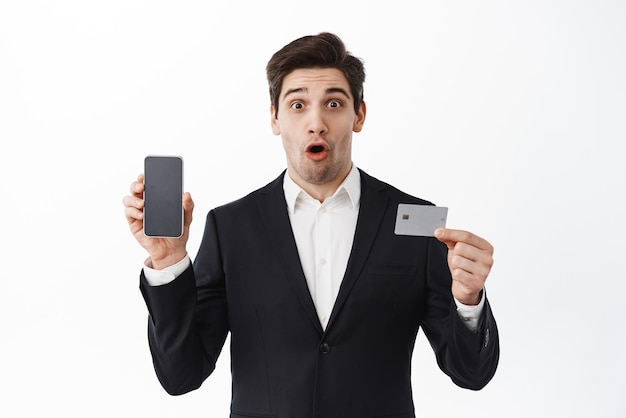 Free Photo impressed entrepreneur showing phone and credit card gasping amazed standing over white background in black suit