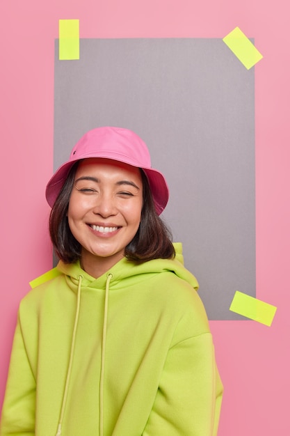 Free Photo indoor shot of happy cute asian teenage girl smiles pleasantly enjoys funny conversation grins at front dressed in casual clothes poses against pink wall plastered paper