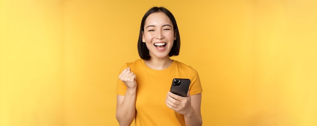 Joyful asian woman celebrating holding mobile phone winning achieve goal on smartphone standing over