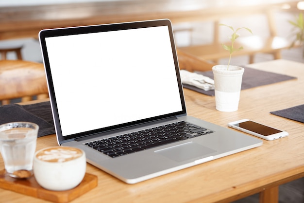 Free photo laptop computer with blank screen. workspace of young freelancer: open notebook pc, cell phone and cappuccino on wooden table