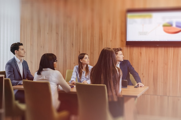 Foto gratuita una grande squadra di persone sta lavorando a un tavolo per laptop, tablet e documenti, sullo sfondo un grande televisore su una parete di legno