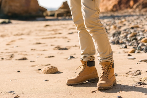 Free photo legs in boots footwear of young hipster man traveling with backpack in autumn sea coast wearing warm jacket and hat