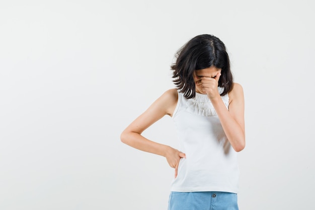 Foto gratuita bambina che strofina gli occhi e il naso in camicetta bianca, pantaloncini e sembra triste, vista frontale.