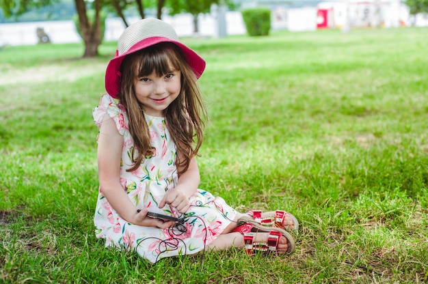 Free photo little girl sitting on the grass looking at a mobile with headphones and with a hat