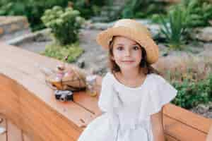 Free photo little tanned lady in vintage straw hat sitting on wooden bench with basket for picnic and camera. outdoor portrait of dark-eyed girl wears white dress posing