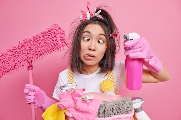 Foto gratuita l'uomo tira fuori la lingua incrocia gli occhi fa una smorfia stupida mentre pulisce la casa tiene il detersivo per la pulizia e il mop si trova vicino alla pila di biancheria posa sul rosa