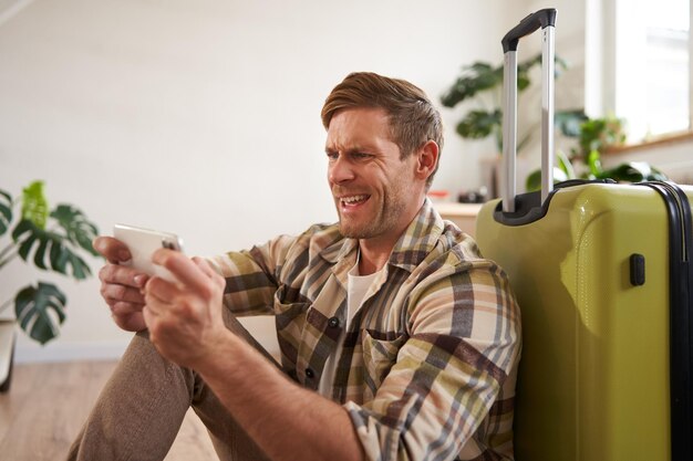 Free photo man with shocked face sitting on floor with suitcase looking worried and tensed on mobile phone