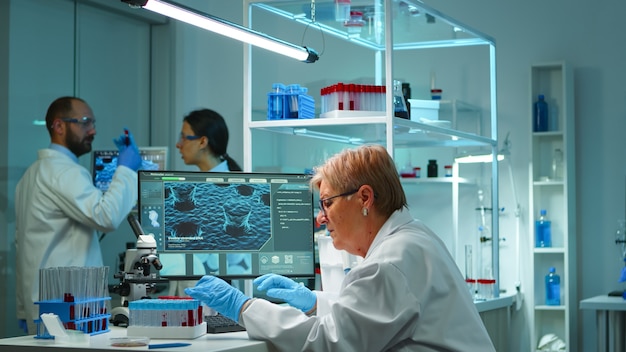 Free photo microchemist doing research at night checking blood liquid in test tube typing results on pc in modern equipped lab. examining virus evolution using high tech for vaccine development against covid19