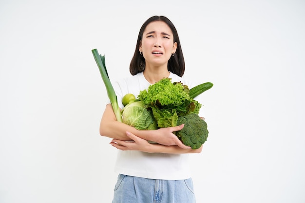 Free photo miserable asian woman holds green vegetables and complains at het diet looks at calories white backg