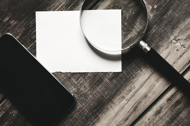 Free photo mobile smartphone, magnifier and sheet of white paper isolated on black farm wooden table mysterious detective game concept. closeup side view