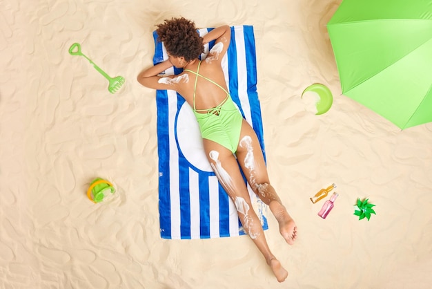 Free Photo overhead shot of relaxed slim woman dressed in green swimsuit lies on stomach sunbathes at beach applies skin care cream has lazy day enjoys summer vacation people leisure relaxation concept