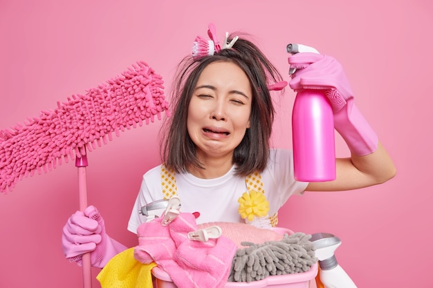 Foto gratuita la foto di una casalinga stressata che piange dalla disperazione stufo della pulizia della casa tiene la scopa e il detersivo ha un'espressione triste che funziona per la casa isolata su sfondo rosa. detergente per la fatica.