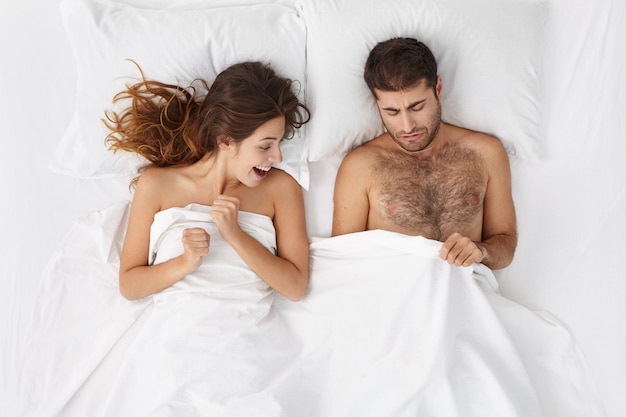 Free photo picture of adult european bearded man and excited woman lying in bed and peeping under white blanket