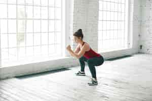 Free photo picture of strong sporty girl wearing stylish tank top, sneakers and leggings doing squats on wooden floor at gym center against large windows