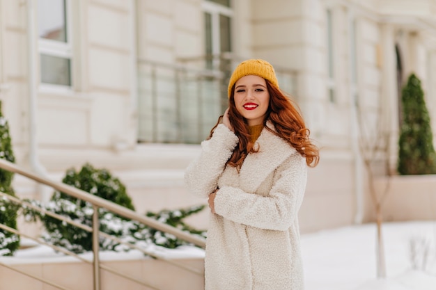 Pleasant caucasian woman in white coat enjoying weekend walk. Outdoor portrait of stylish ginger girl in winter outfit.