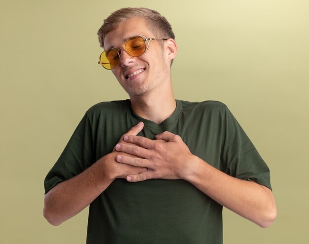 Pleased with closed eyes young handsome guy wearing green shirt with glasses putting hands on heart isolated on olive green wall