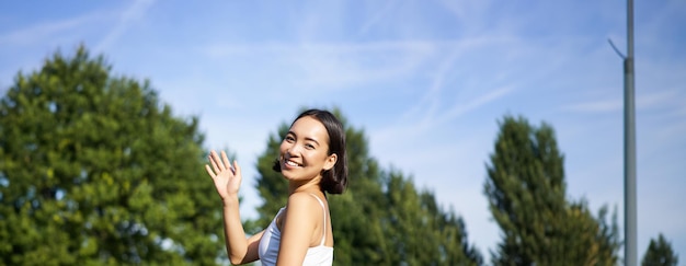 Foto gratuita ritratto di una ragazza asiatica in classe di fitness nel parco seduta su un tappetino di gomma e agitando la mano alla telecamera