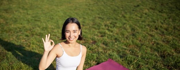 Foto gratuita ritratto di una donna asiatica seduta su un tappetino di gomma che medita mostrando un segno okay e guardando la telecamera