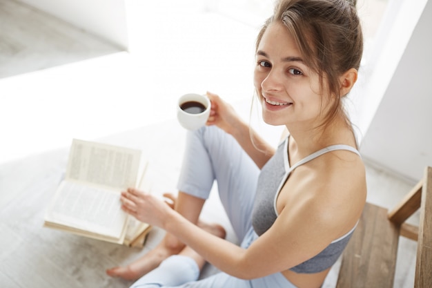 Free photo portrait of beautiful happy woman smiling holding cup of coffee sitting on floor with book.