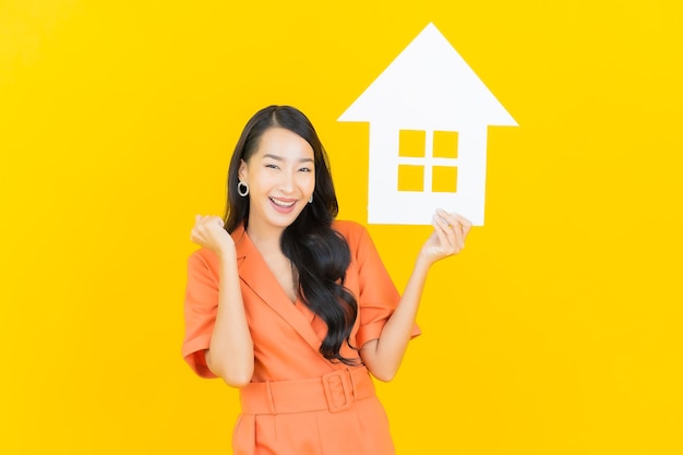 Free photo portrait beautiful young asian woman smile with home sign paper board on yellow