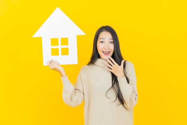 Free photo portrait beautiful young asian woman with house or home paper sign on yellow wall