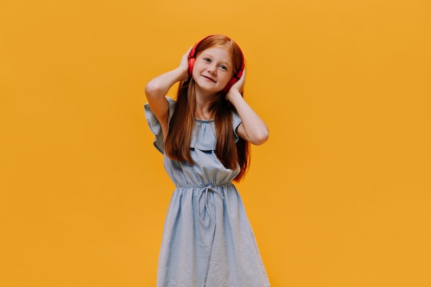 Free photo portrait of charming little girl listening to music beautiful redhead child in blue dress poses in red headphones on orange background