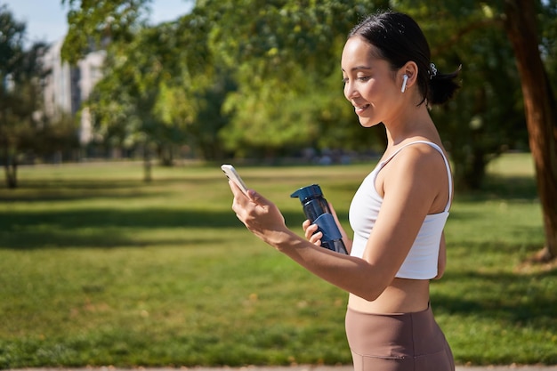 Free photo portrait of fitness girl runner drinking water looking at smartphone standing in park in middle of w