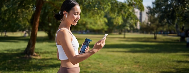 Free photo portrait of fitness girl runner drinking water looking at smartphone standing in park in middle of