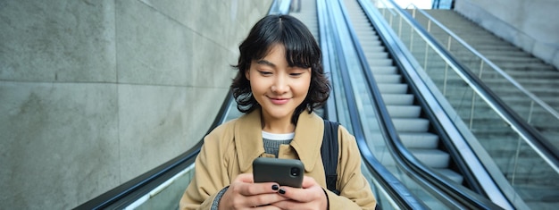 無料写真 美しいアジアの女子学生の肖像画が街のエスカレーターを下り、携帯電話の使用状況を確認する