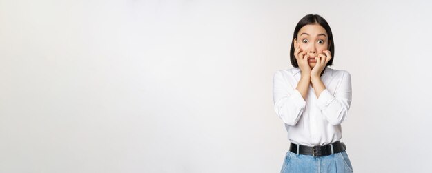 Portrait of scared asian woman watching smth scary biting fingers on hands and looking at camera frightened standing over white background