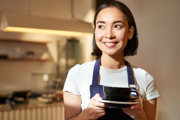 Free photo portrait of smiling asian barista girl in uniform serving coffee waitress holding cup of drink working in cafe