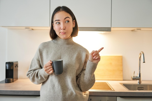 Free photo portrait of smiling asian girl standing in kitchen drinking coffee from cup and pointing at banner s