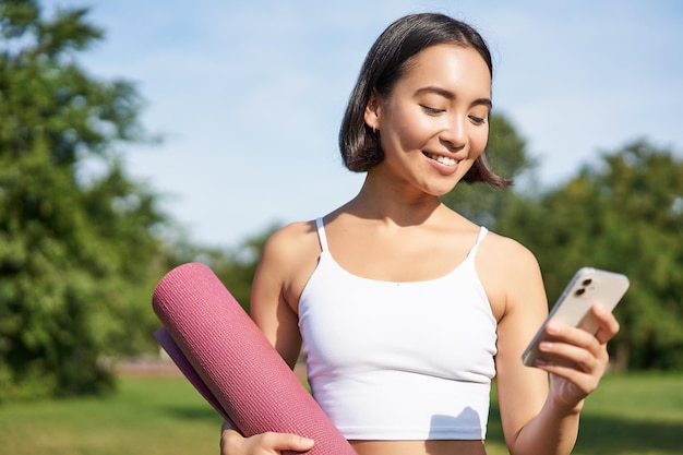 Free photo portrait of smiling asian woman with yoga mat looking at her smartphone and reading on application standing in park wearing sport uniform