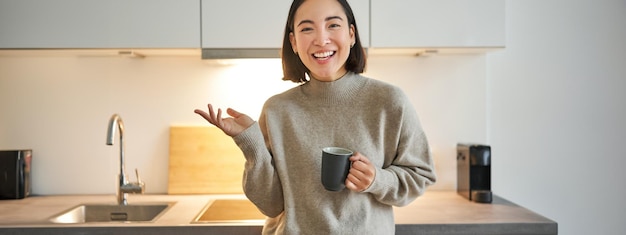 Free photo portrait of stylish asian woman standing in kitchen with mug drinking coffee explaining smth showing